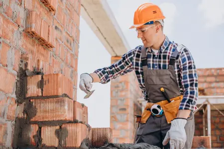 Junger Handwerker in Schutzhelm und Handschuhen baut eine Backsteinmauer.